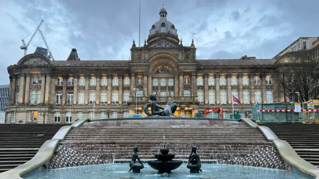A large council building which has dozens of windows and  a first floor balcony. It has a dome tower on the top. In front of it is a large water feature surrounded by steps. There is a statue at the top of the steps in the water, and two smaller statues and a water feature further down