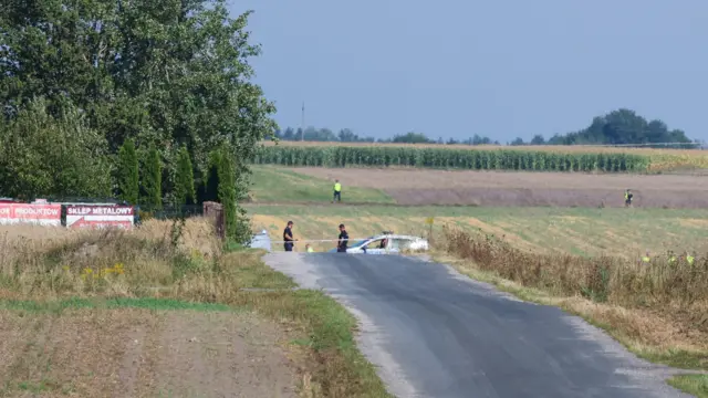 A far-away shot of a field, with a lane leading up to it and a few uniformed officers - some in high-vis - inspecting the site.