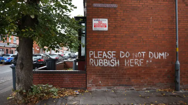 A red brick wall on a street corner with words spray painted on it in white. The writing reads "please do not dump rubbish here". There is a sign on the wall that says "no dumping"