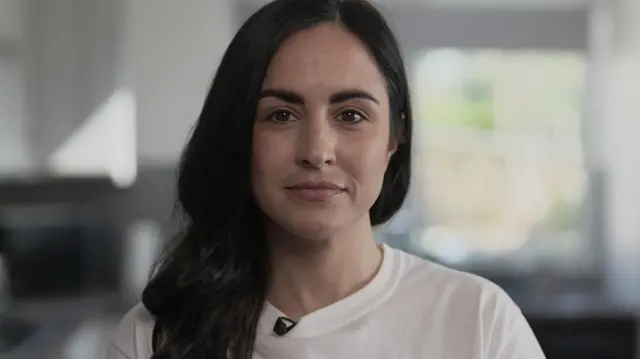 Leanne Lucas with long dark brown hair standing in a kitchen wearing a white t-shirt
