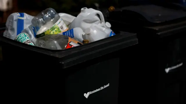 black bin with a sticker on it that reads "birmingham city council". It is full to the brim with recyclable materials including plastic bottles and milk cartons