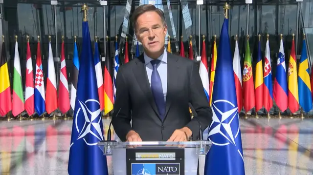 Man in suit and glasses speaking ito mic in front of various country flags