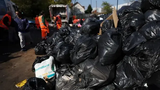 A huge pile of black bin bags in the foreground. IN the background is a white bin lorry with refuse workers standing nearby in orange hi-vis