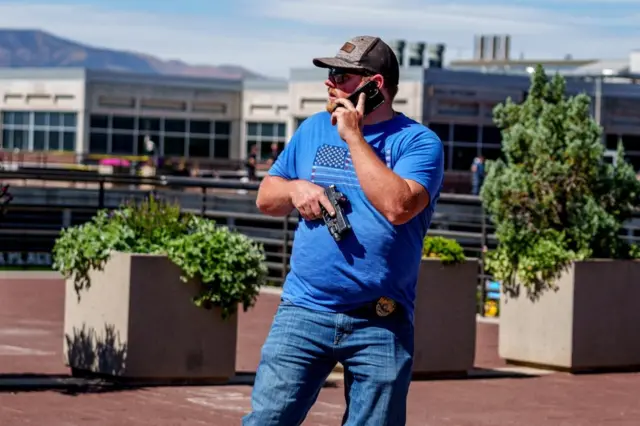 An officer is seen in plainclothes, with a handgun drawn and speaking on the phone