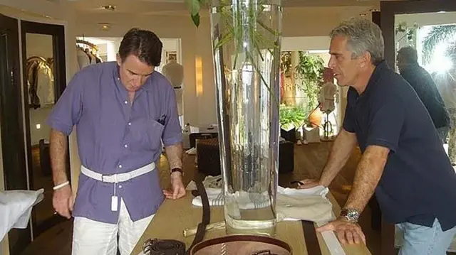 Mandelson looks down at a white belt he is wearing which has a tag on it while on the other side of a table stood across from him is Epstein, leaning on the table, watching Mandelson