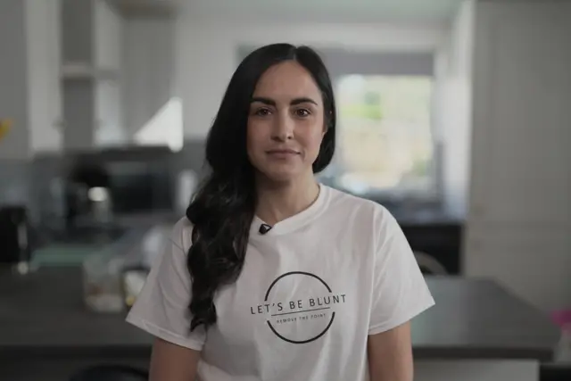 Leanne Lucas, who has long dark brown hair, stands in a kitchen looking into the camera while wearing a white t-shirt