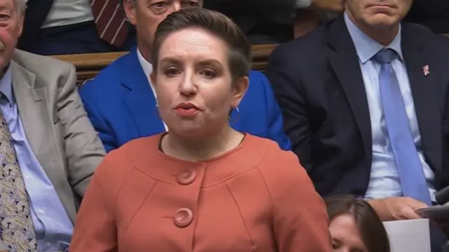 Headshot of Denyer who stands up and speaks. She wears an orange buttoned up coat and people sit on benches behind her