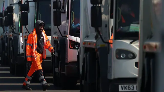 A man with his hood up is wearing an orange hi-vis jacket. He has a cigarette in his hand and is walking towards a line of parked white bin lorries