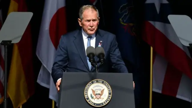 George W Bush speaks with his hands on a lectern while standing in front of national flags