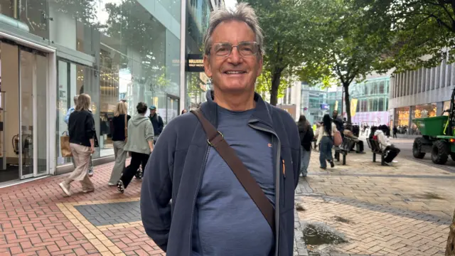 A man with long brown hair is standing in the street. He is wearing glasses and a navy hoodie with a blue top. He is also wearing a brown cross-body bag