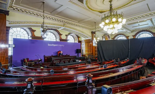 Sir Adrian Fulford sits at an ornate mahogany bench in a council chamber at Liverpool town hall. He is faced by several rows of curved benches. Behind him is a purple screen with 'Southport Inquiry' printed on it in white lettering.