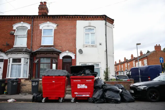 Two red Biffa branded general waste bins outside terraced housing. They are filed and overflowing with black bin bags. There is a pile of black binbags on the floor, with a car parked behind them