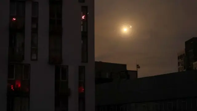 A fireball appears in the night sky near a residential building.