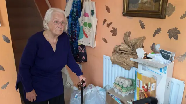 Ms Boyce standing in her hallway. She is leaning on her walking stick. It is painted in orange with a leafy pattern. She is standing next to a pile of recycling. We can see cardboard boxes, plastics and some bottles by her feet.