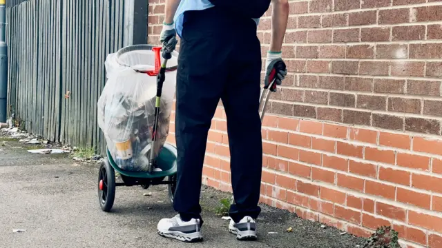 The bottom half of a man facing away from the camera. He is standing next to a brick wall and is pushing a trolley along that has a binbag attached to it, filled with rubbish. He is wearing gloves and has a grabber in his hand