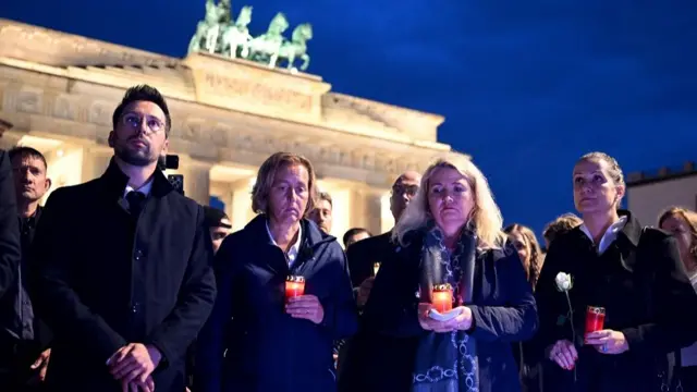 Four people stand in front of a crowd, holding candles and one of them has a white rose. Their faces are sombre