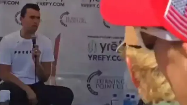 Charlie Kirk, speaking into microphone wearing white t-shirt, sitting on a chair.