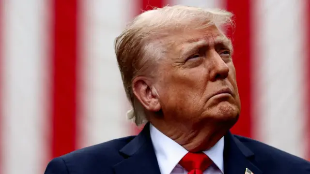 Donald Trump looks to the side, wearing a suit and red tie, with an American flag draped in the background.