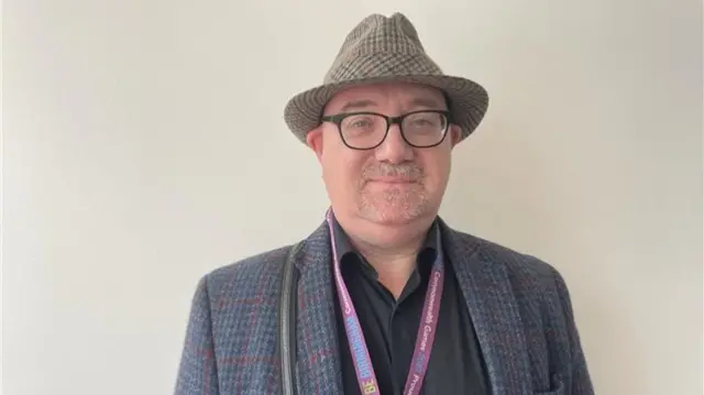 A man with a beige, black and red striped hat on. He is wearing black glasses and has a blue and red striped blazer on, with a black shirt and purple lanyard around his neck.