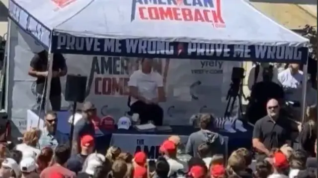 A large crowd of people look at Charlie Kirk who is sitting under a white marquee.