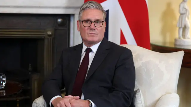 The UK Prime Minister Sir Keir Starmer sat in a white chair. A Union Jack flag hangs behind him.