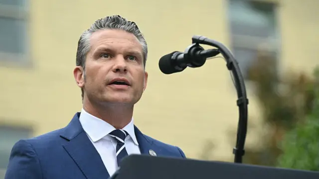 Hegseth wears a suit and speaks behind a podium.