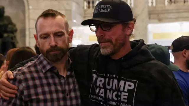 Two men looking down, with one putting his hand over the other's shoulder. One is wearing a Trump Vance hoodie, and the other is in a flannel shirt