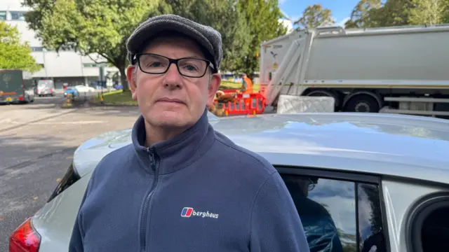 A man wearing a grey flat cap and a blue fleece looks towards the camera. He is standing in front of a car