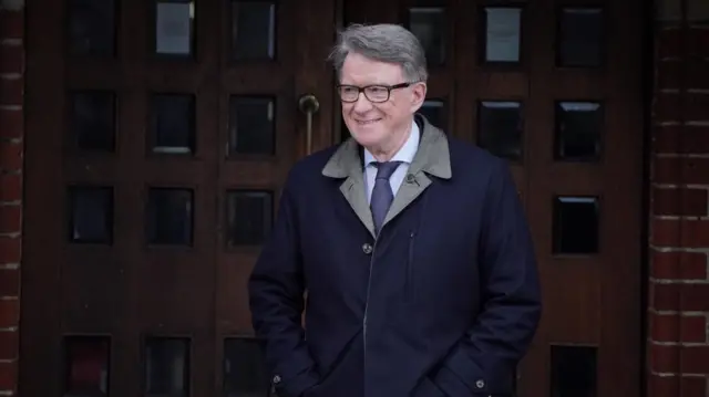 Man in tie and jacket smiles whilst walking in front of a brown door