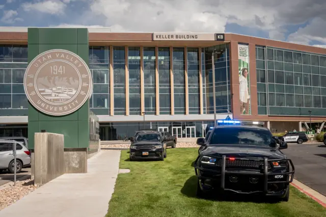 The exterior of the Keller building at the Utah Valley University during daylight. A black police vehicle is parked on a grassy area outside the building, with blue lights flashing. Three other police vehicles are parked outside the building.