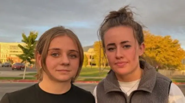 Two young women stand side by side in front of the camera - the one on the left wears a black tee shirt, and the one on the right wears a fleece vest over a short-sleeved white tee shirt
