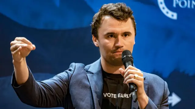 Charlie Kirk, holding a microphone and gesturing with hand, wearing a "Vote Early" t-shirt and blue blazer
