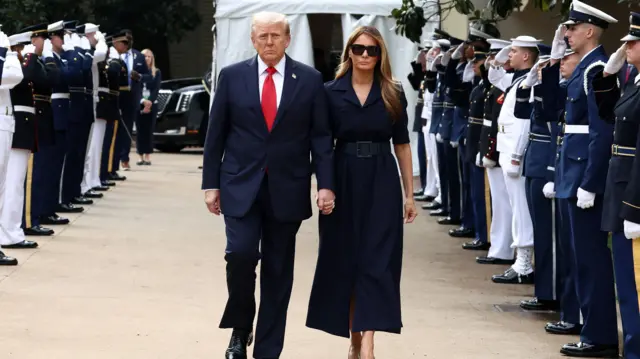President Trump and Melania walk towards the camera dressed in dark clothing and holding hands. Military personnel line their path and salute.