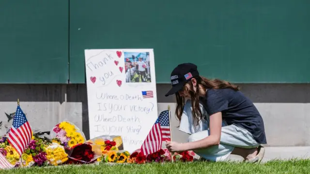 A girl wearing a MAGA hat kneels down at a makeshift memorial site at the Utah Valley University. There are flowers and USA flags at the site, with a sign of Kirk reading: Thank you, Where, O Death, Is your victory? Where, O Death, Is your sting?