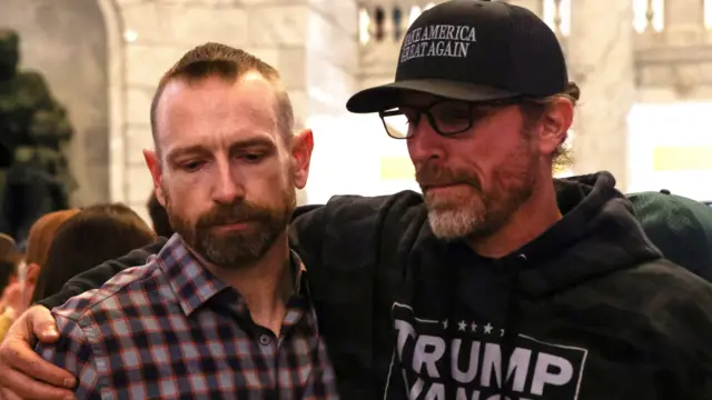A man in a black MAGA hat and a Trump-Vance hoodie puts his arm round another man in a chequered shirt