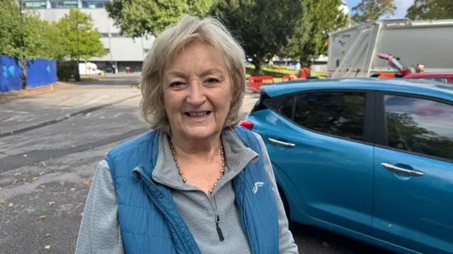 A woman with short blonde hair is wearing a light grey top and a blue gilet. she is standing next to a blue car