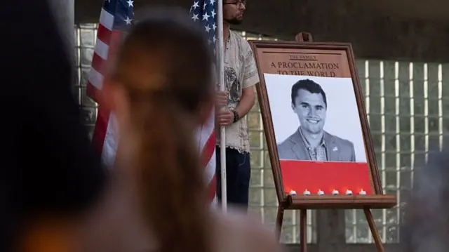 Blurred backview of people attending a vigil. On an easel is Charlie Kirk's photo in black and white.