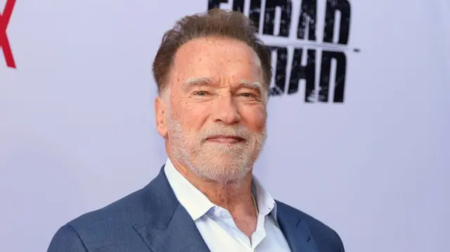 Arnold Schwarzenegger, smiling at camera with grey stubble, wearing grey blue suit