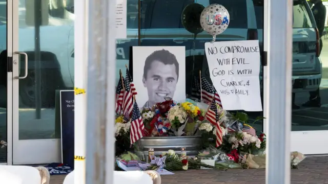 A small makeshift memorial with USA flags, and flowers, with a photo of Charlie Kirk's face