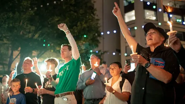 A vigil being held in memory of Charlie Kirk. Two people, a man in a green t-shirt and woman in a black shirt, stand at the front holding candles, they have their fists clenched and raised in the air.
