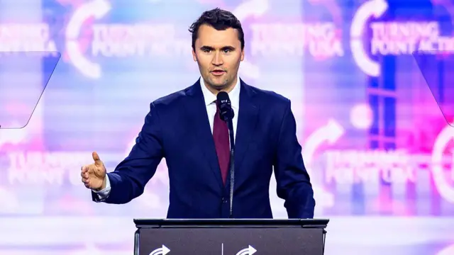 Charlie Kirk, wearing blue suit, speaking at a podium with Turning Point USA branding