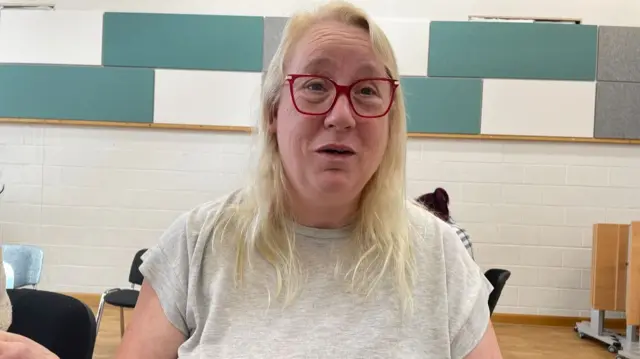 A woman with blonde shoulder length hair wearing red glasses and a grey top