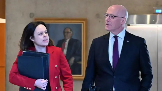 a woman with shoulder-length black hair and wearing a red jacket is carrying black folders. She is stranding next to a bald man with glasses who is wearing a dark suit, white shirt and purble tie