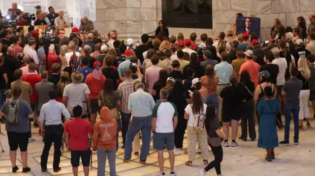 A large group of people gather in front of a white-bricked wall in memory of Charlie Kirk. At the front of the group a woman speaks into a microphone, and a portrait of Kirk is displayed.