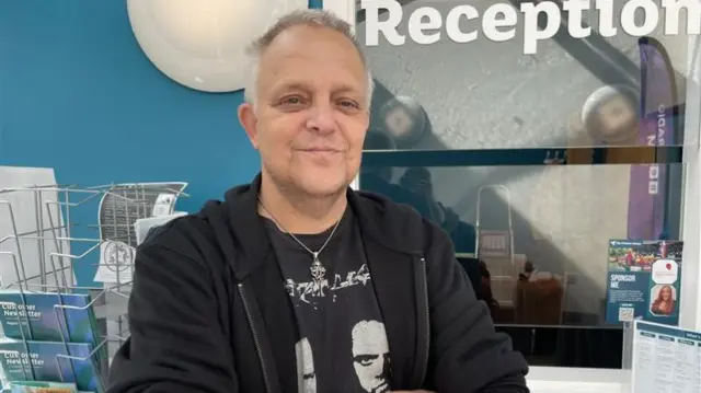 A man with short white and grey hair. He is wearing a silver necklace, black zip hoodie and a black tshirt.