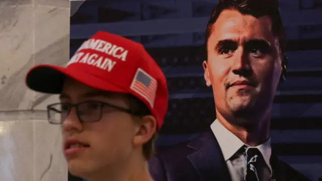 A bespectacled teenager with a red Make America Great Hat. Behind him is a large photo of Charlie Kirk.