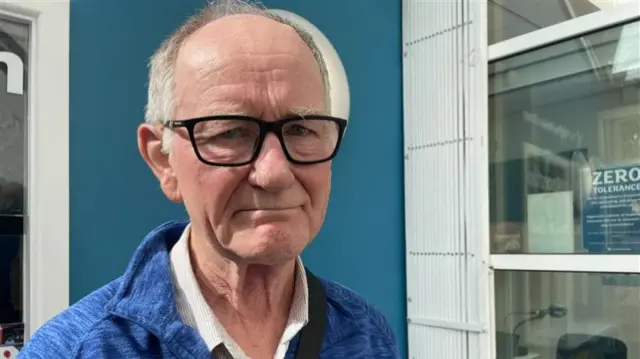 A man with short grey hair and black glasses. He is wearing a dark blue top with a white shirt underneath