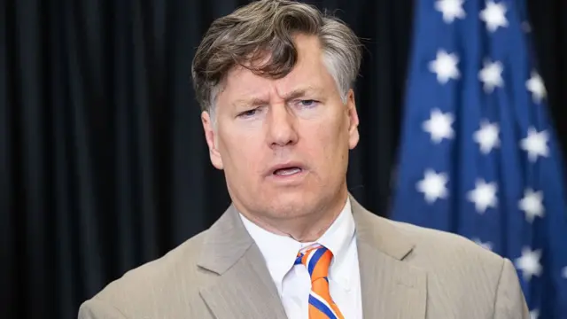 Christopher Landau speaks wearing a brown suit and orange tie.