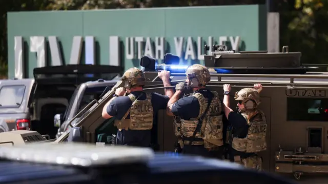 Police officers, wearing camouflage guests, crouch next to a police vehicle. A sign reads Utah Valley University behind them