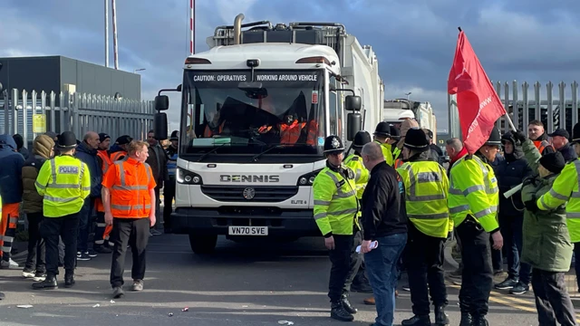 A white bin lorry exits a depot  as union members picket either side of the road as police officers in high-vis jackets try to control the picketers